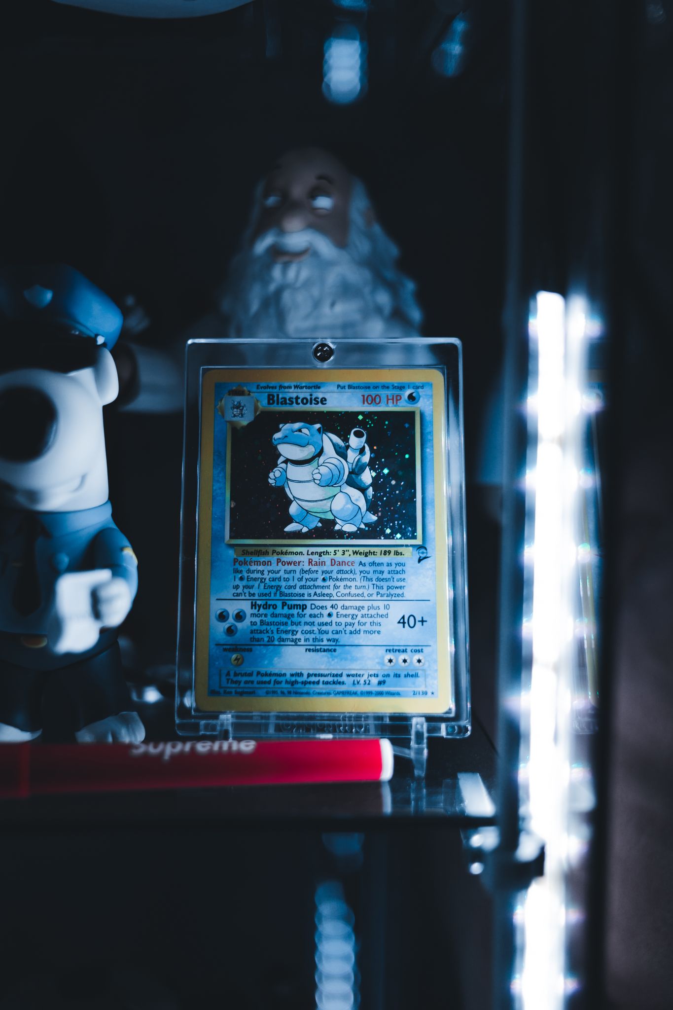 Blastoise Pokémon card in a protective case on display shelf with collectibles and ambient lighting.
